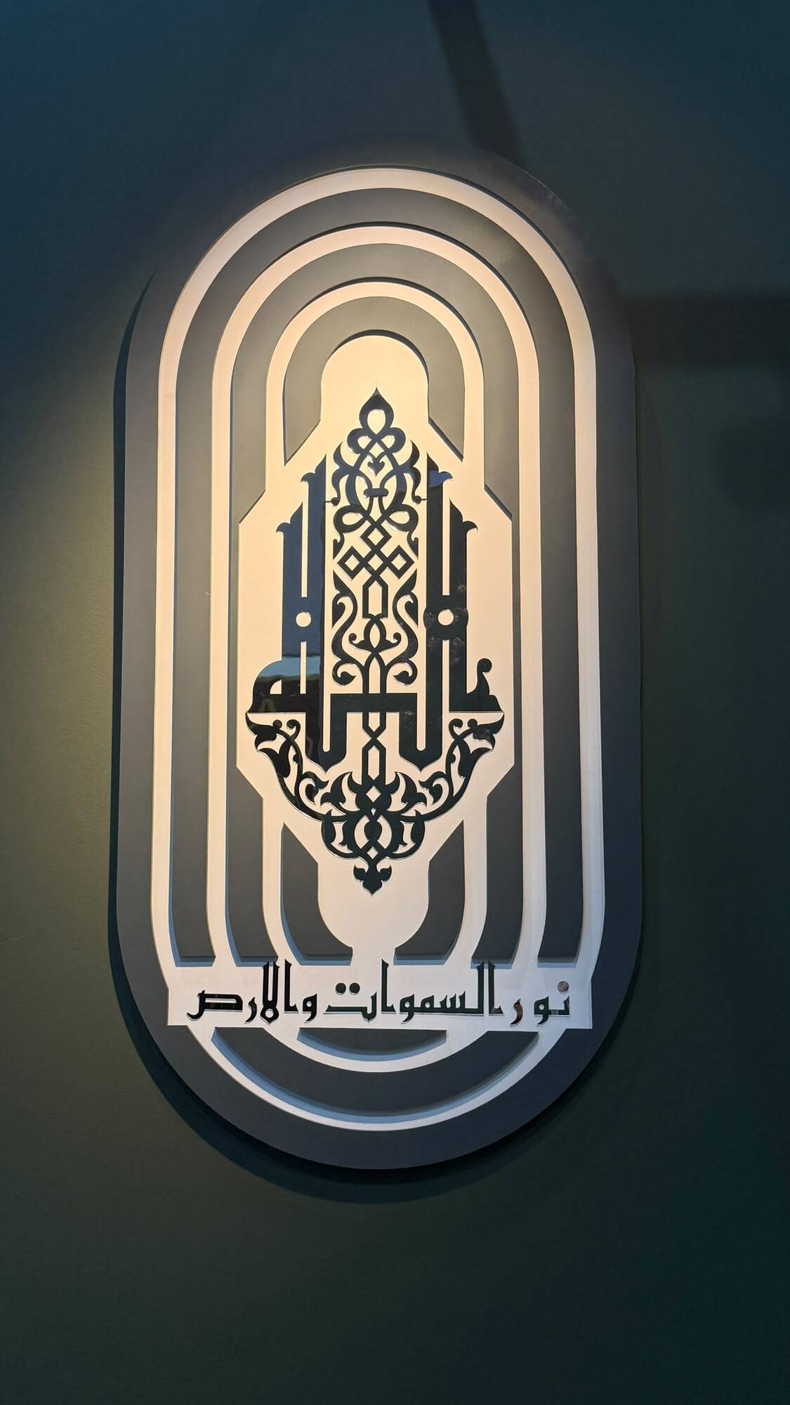 Backlit layered oval wall panel with gold calligraphy glowing on a dark wall